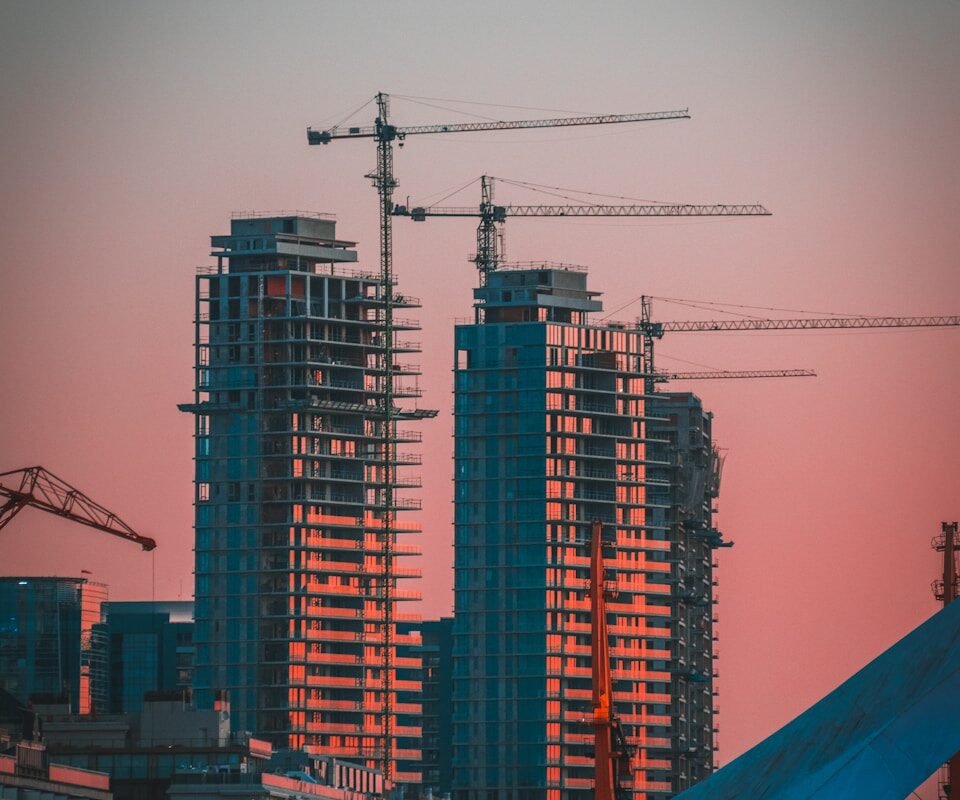 Höghus under byggnation med byggkranar i stadssiluett vid solnedgång med rosa himmel
