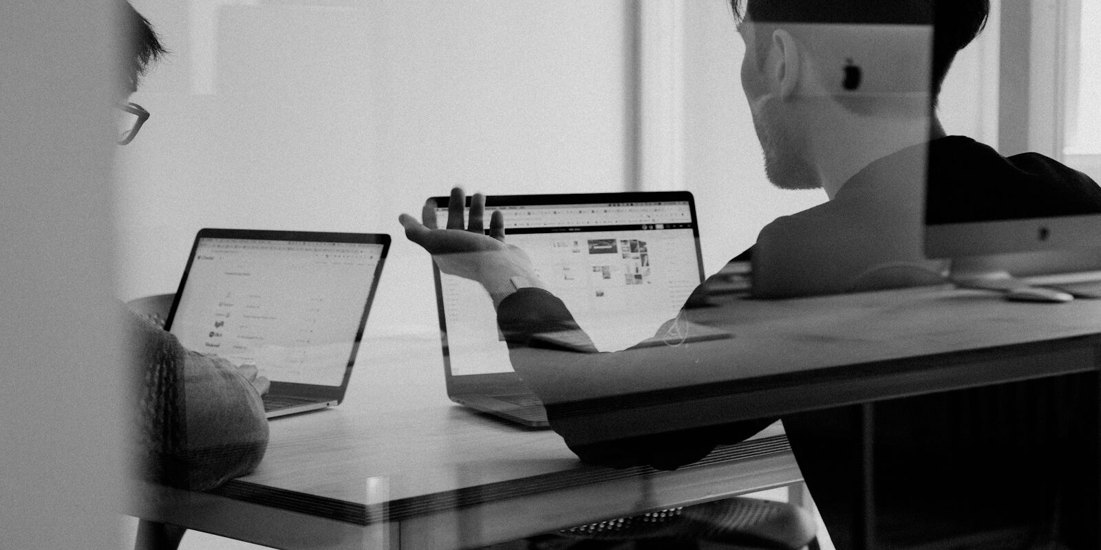 Två kollegor i mötesrum diskuterar arbete vid bord med öppna bärbara datorer, svartvit bild genom glas.